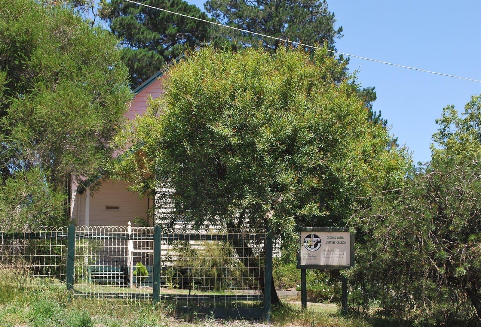 A small brick church half hidden by a tree, behind a wire fence