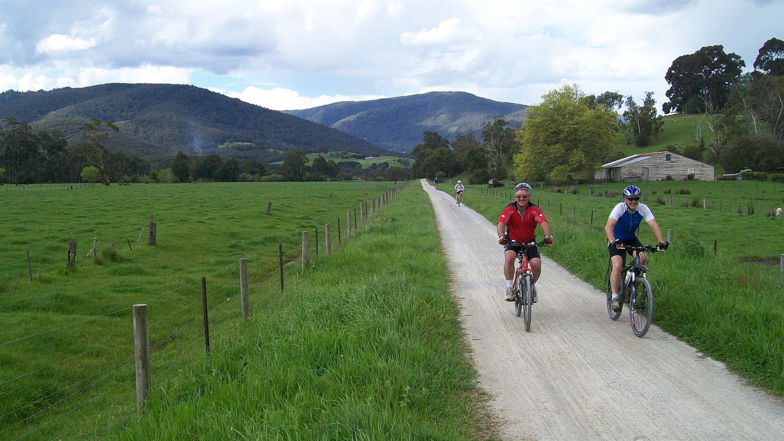 Two cyclists on a gravel trail between paddocks, with ranges behind