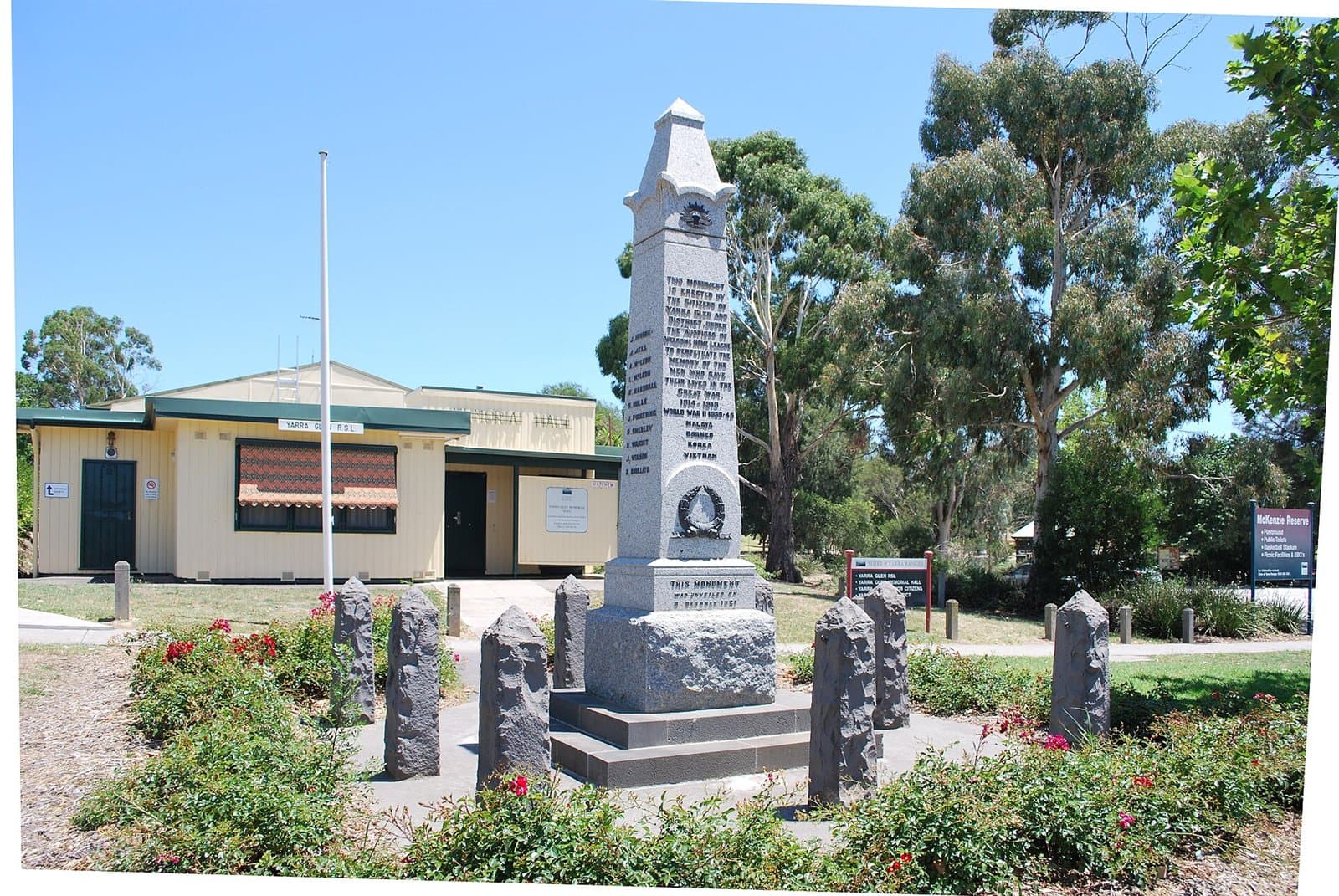 A stone war memorial in a garden bed, with a flagpole and a hall behind